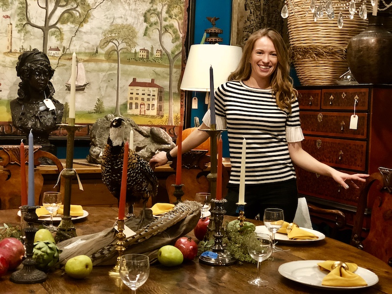Designer, Diane Rath, stands near one of her unique Thanksgiving tablescape designs featuring bird and fowls, vintage candlesticks and fruit